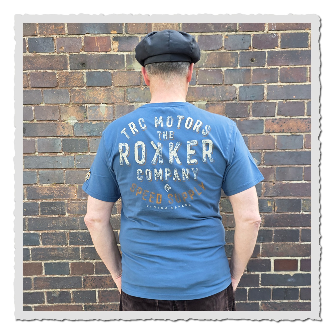 Mann von hinten in blauem T-Shirt mit großem Rückenprint, dunkler Kappe, vor mehrfarbiger Backsteinwand.